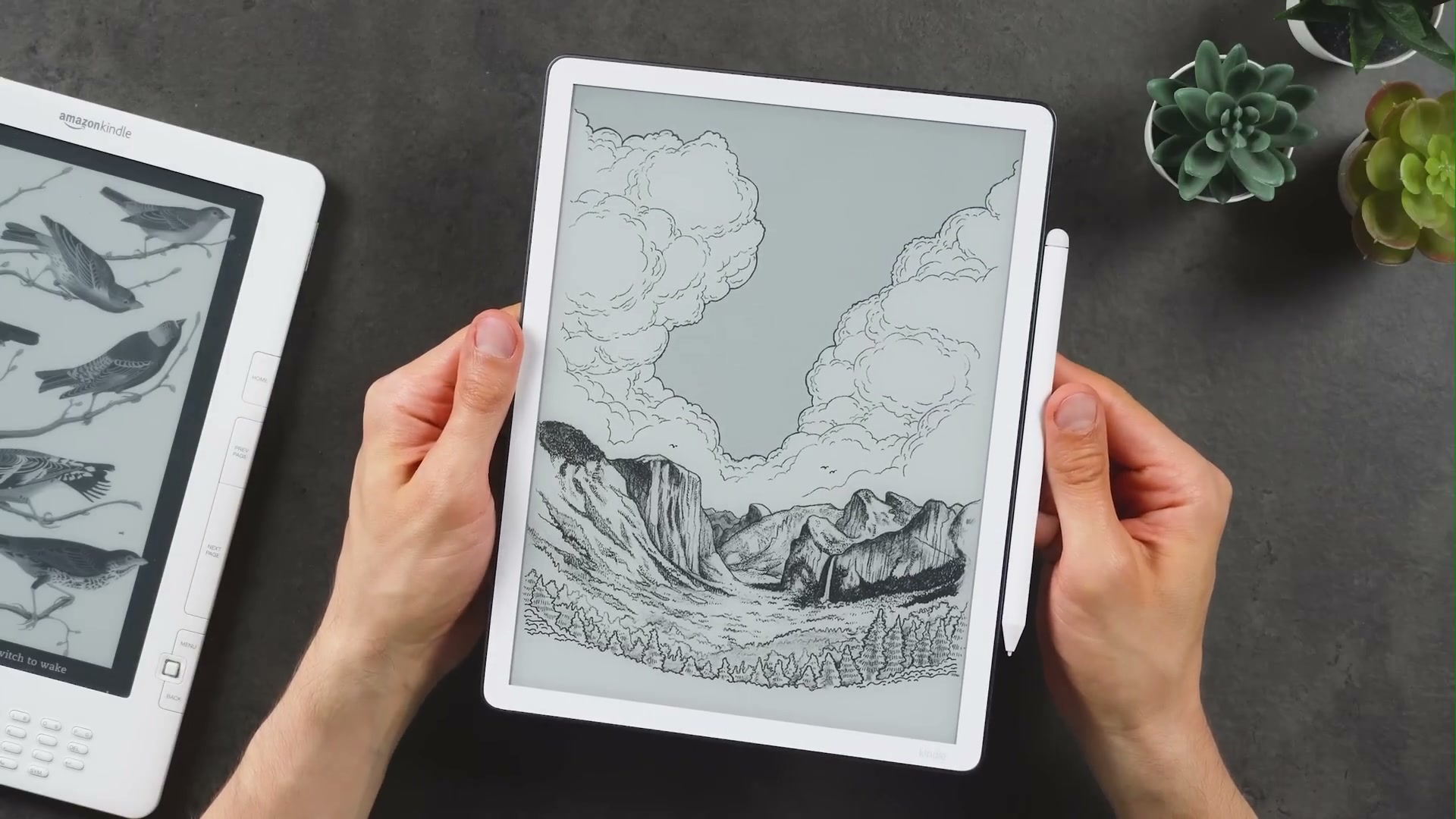 Neuer Kindle Scribe mit weißem Rahmen, Bergmotiv und rechts befestigtem Stift in beiden Händen.