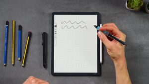 Scribe Colorsoft mit Schreiblinien und einem anderen Stift in der Hand; der weiße Kindle-Stift haftet rechts.