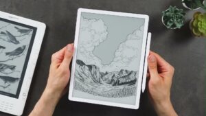 Neuer Kindle Scribe mit weißem Rahmen, Bergmotiv und rechts befestigtem Stift in beiden Händen.