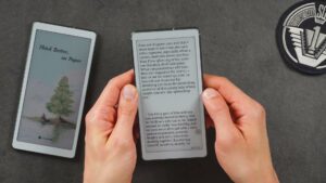 Schwarzweiß-AiPaper Reader in beiden Händen mit einer Buchseite; der Reader C liegt links im Hintergrund.